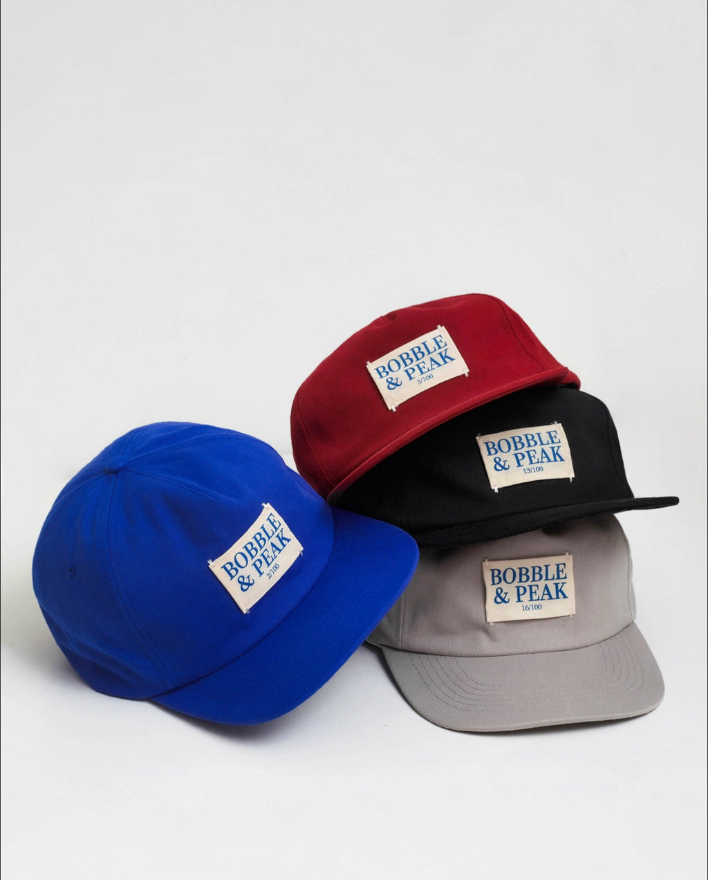Four baseball caps in blue, red, black, and gray with 'Bobble & Peak' label on a light gray background